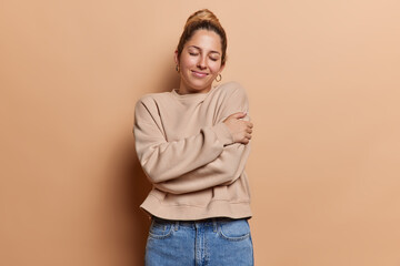 Photo of pleasant looking tender woman European has eyes closed embraces herself dressed in casual jumper and jeans stands against brown studio background enjoys herself. I love myself concept