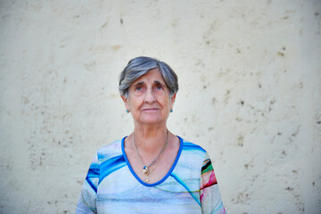 Octogenarian woman with short gray hair, a blouse in shades of blue and pendants and jewelery earrings poses in front of a rough concrete wall painted off-white