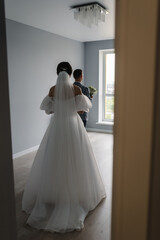 Fototapeta premium Bride goes back to groom, surprise at home. Handsome groom with bouquet flowers in white room. Groom is waiting for the bride. Back view. Happy wedding day of marriage. The first meeting is newlyweds.