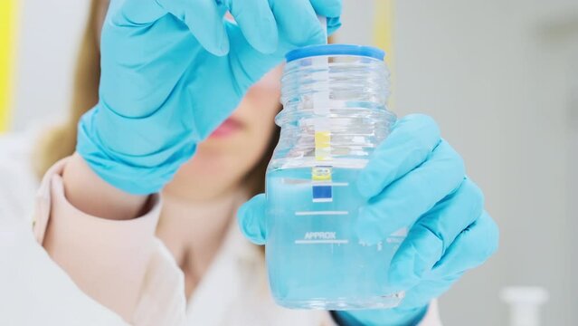 Woman chemist in rubber gloves and lab coat tests solution for the pH using indicator paper. 