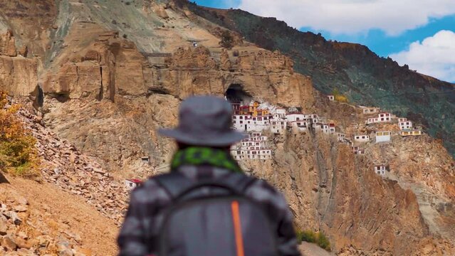 Defocused Indian male tourist wearing hat walking in front of the Phugtal Monastery at Zanskar in Ladakh, India. Traveler trekking towards the Phugtal monastery made inside cave on a mountain.