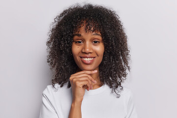 Portrait of cheerful young woman with ark curly hair touches chin smiles toothily being in good mood dressed in casual t shirt isolated over white background. People and happy emotions concept
