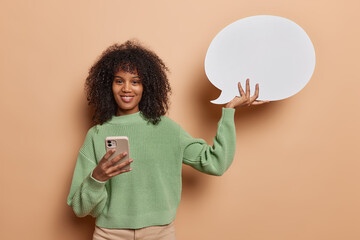Horizontal shot of pretty cheerful African woman holds smartphone within white communication bubble...