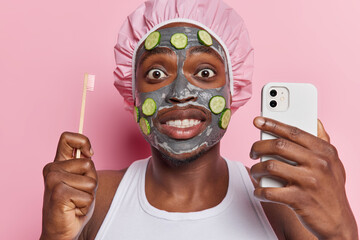 Facial treatments and dental care concept. Shocked African man applies clay nourishing mask and cucumber slices holds smartphone and toothbrush wears white t shirt and bath hat isolated on pink wall