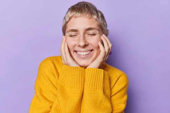 Portrait of cheerful woman with blonde short hair keeps eyes closed touches cheeks smiles gently expresses happy sincere emotions wears yellow jumper isolated over purple wall. People and positiveness