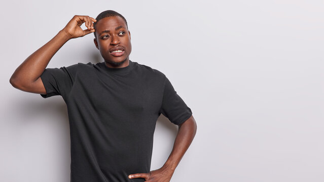 Thoughtful dark skinned man being deep in thoughts scratches head concentrated aside tries to solve problem wears casual black t shirt isolated over white background. Uncertain black guy poses indoors