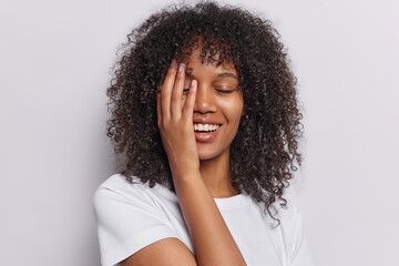 Curly haired woman radiates positivity as she covers half her face with her palm eyes closed in blissful contentment dressed in casual tshirt isolated on white background. People and happiness concept
