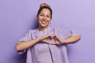 Horizontal shot of pretty young European woman makes heart hand gesture demonstrates love sign...