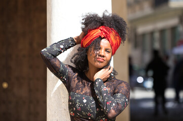 Portrait of a young, beautiful black woman with afro hair and black dress with flowers, wearing a red scarf in her hair. The woman is happy and laughing and having fun leaning on a marble column.