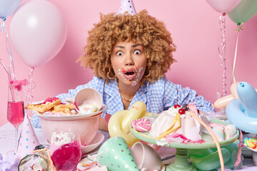 Obraz na płótnie Canvas Startled young woman with curly hair wears cone hat and pajamas poses near festive table with desserts celebrated birthday isolated over pink background. People celebration and partying concept
