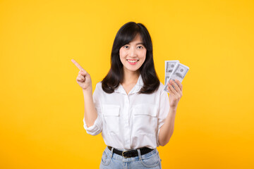 Young happy woman of Asian ethnicity wear white shirt and denim jean holding cash money in dollar and pointing finger to free copy space against yellow background. Investment and financial concept.