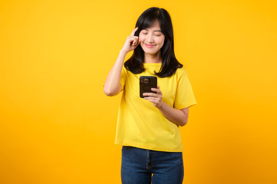 Experience Range Of Emotions With Expressive Portrait. Young Asian Woman Wearing Yellow T-shirt And Denim Jeans Showcases Doubtful Expression While Using Smartphone. Diverse Emotions And Tech Usage.