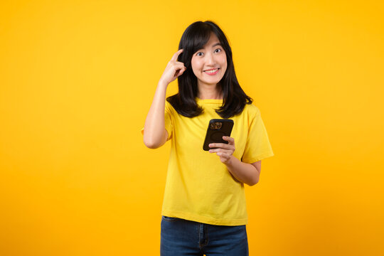 Experience Range Of Emotions With Expressive Portrait. Young Asian Woman Wearing Yellow T-shirt And Denim Jeans Showcases Doubtful Expression While Using Smartphone. Diverse Emotions And Tech Usage.