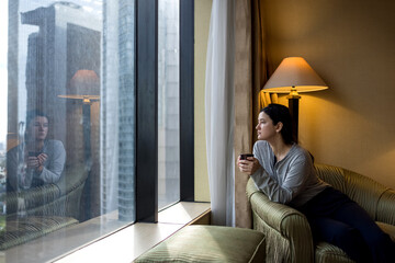 Young woman holding coffee cup, wearing pajama and looking at cityscape through the window in luxury penthouse apartment or hotel room - 619149375