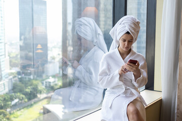 Young woman in bathrobe and towel on the head using smartphone, sitting in luxury penthouse apartment or hotel room with city skyscraper view. Businesswoman on vacation or holidays