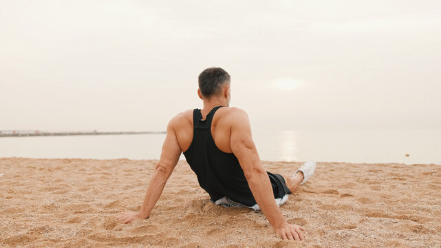 Middle-aged athletic man sits on the beach after workout and looks at the sea, Back view