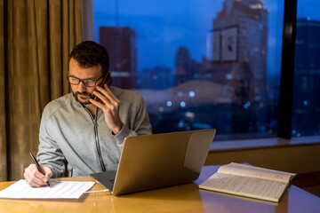 Handsome man using laptop and mobile phone in penthouse or luxury hotel room doing paper work report. Businessman in skyscraper apartment against night city background working late at nigh.