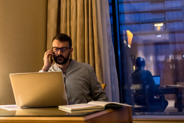 Handsome man using laptop and mobile phone in penthouse or luxury hotel room doing paper work...