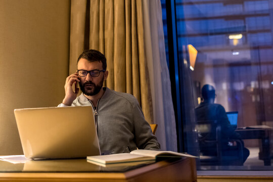 Handsome man using laptop and mobile phone in penthouse or luxury hotel room doing paper work report. Businessman in skyscraper apartment against night city background working late at nigh.