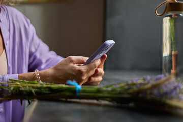 Young woman in violet clothes sitting behind a table in a cafe and typing a message on a smartphone. Unrecognizable female person in stylish purple outfit texting online with a modern mobile phone