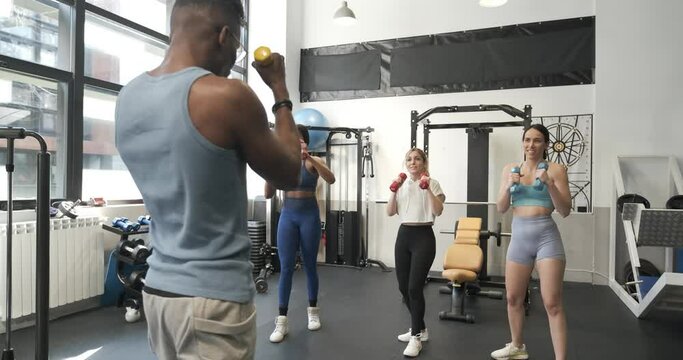 Happy diverse women doing workout with dumbbells and trainer in gym