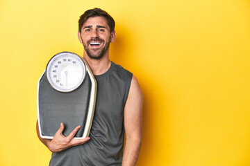 Athletic man with scale, on a yellow studio backdrop happy, smiling and cheerful.