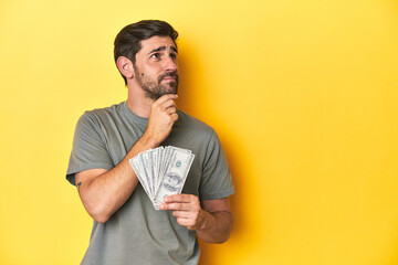Caucasian man holding dollars, yellow studio shot looking sideways with doubtful and skeptical...
