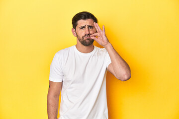 Caucasian man in white t-shirt on yellow studio background with fingers on lips keeping a secret.