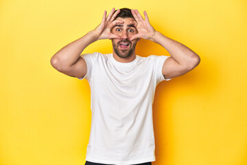 Fototapeta na wymiar Caucasian man in white t-shirt on yellow studio background keeping eyes opened to find a success opportunity.
