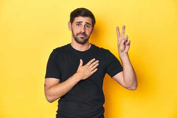 Caucasian man in black t-shirt, yellow studio backdrop taking an oath, putting hand on chest.