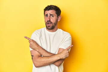 Caucasian man in white t-shirt on yellow studio background points sideways, is trying to choose...
