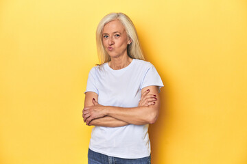 Middle-aged Caucasian woman in white t-shirt, yellow studio frowning face in displeasure, keeps arms folded.