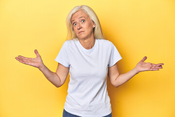 Middle-aged Caucasian woman in white t-shirt, yellow studio doubting and shrugging shoulders in...
