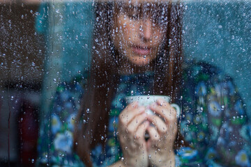 mujer sujetando con las manos taza de café detrás de cristal con gotas con vestido azul