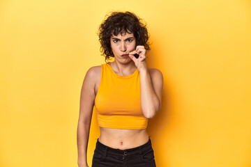 Curly-haired Caucasian woman in yellow top with fingers on lips keeping a secret.