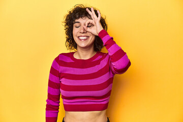 Caucasian curly-haired woman in pink striped-top excited keeping ok gesture on eye.
