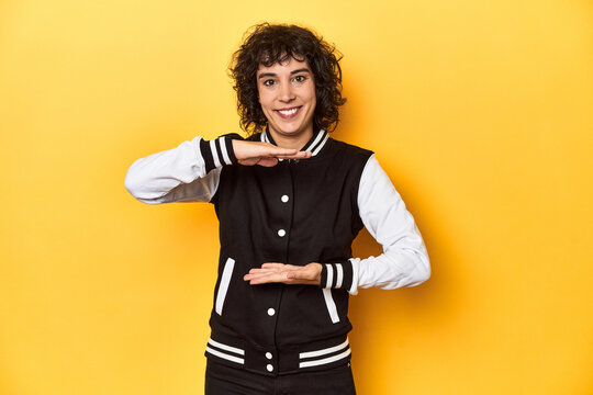 Curly-haired Caucasian woman in baseball jacket holding something with both hands, product presentation.