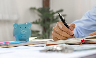 Businessman hand writing income and expenses to account with piggy bank and dollar notes, step up start-up businesses to success, Saving money for future plan and retirement fund concept