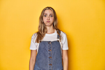 Young blonde Caucasian woman in denim overalls posing on a yellow background, blows cheeks, has...