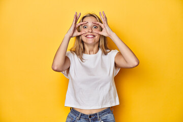 Young blonde Caucasian woman in a white t-shirt on a yellow studio background, keeping eyes opened to find a success opportunity.