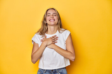 Young blonde Caucasian woman in a white t-shirt on a yellow studio background, laughing keeping...
