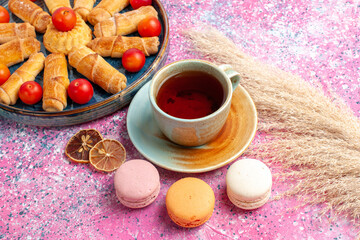 front view sweet delicious bagels inside tray with fresh sour plums french macarons and cup of tea on light-pink desk cake biscuit cookie sweet sugar pie