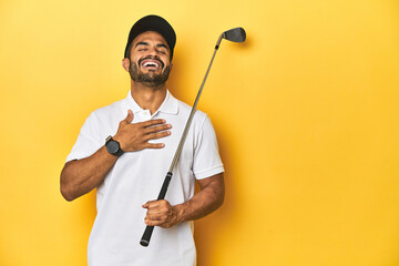Young Latino golfer with club and cap on a yellow studio background, laughs out loudly keeping hand on chest.