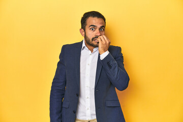 Professional young Latino man in business suit, yellow studio background, biting fingernails, nervous and very anxious.