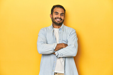 Casual young Latino man against a vibrant yellow studio background, who feels confident, crossing arms with determination.