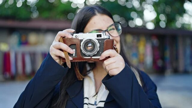 Young hispanic woman tourist wearing backpack taking pictures with camera at street market