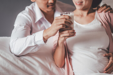 Concept Maternity and Pregnant, Prenatal care and pregnancy. Portrait of Pregnant woman sitting on a white bed Her husband holds a glass of milk and sends her fresh milk in a clear glass for her to dr