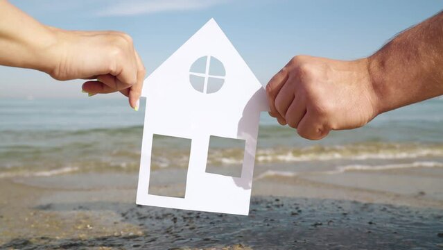 Symbol of home, happiness, Concept of building house for family on sea coast. Dreaming of buying house by sea, family life. Hands of family hold paper house in front of sea, view from window to ocean