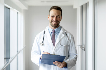  Portrait of a friendly european doctor in work clothes with a stethoscope around his neck, posing with folded hands in the interior of the clinic, looking and smiling at the camera, free space