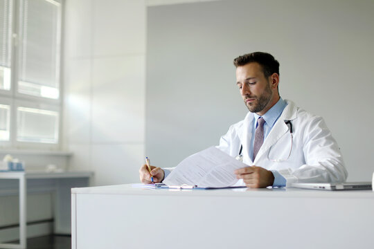 Young male doctor sitting at table in clini office, practitioner writing and working, checking patients results and writing prescriptions, copy space